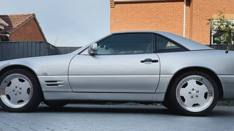 A silver Mercedes-Benz SL R170 coupe parked on a paved driveway in front of a brick house.