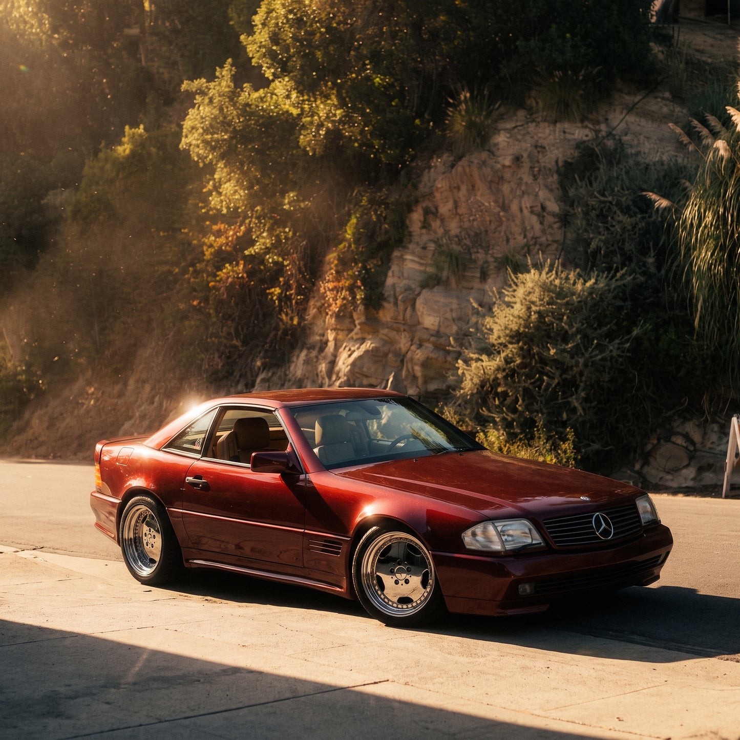 A dark red Mercedes-Benz convertible coupe parked on a sunlit road with a rocky, vegetated hillside in the background.