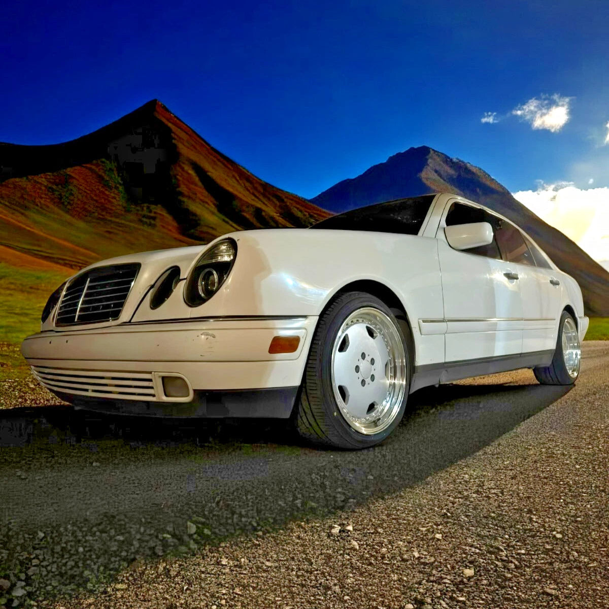 A white Mercedes-Benz sedan parked on a gravel road with mountains and a blue sky in the background.