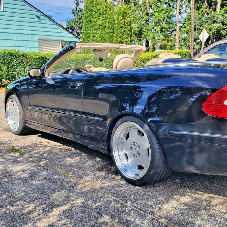 A black convertible car parked outdoors on a paved surface, with the top down and beige seats visible. The photo is taken in a real outdoor setting with trees, a house, and another vehicle in the background.