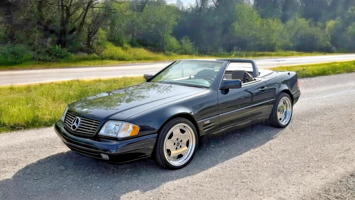A black Mercedes-Benz SL convertible parked on a gravel road outdoors, with grass, trees, and a road visible in the background.