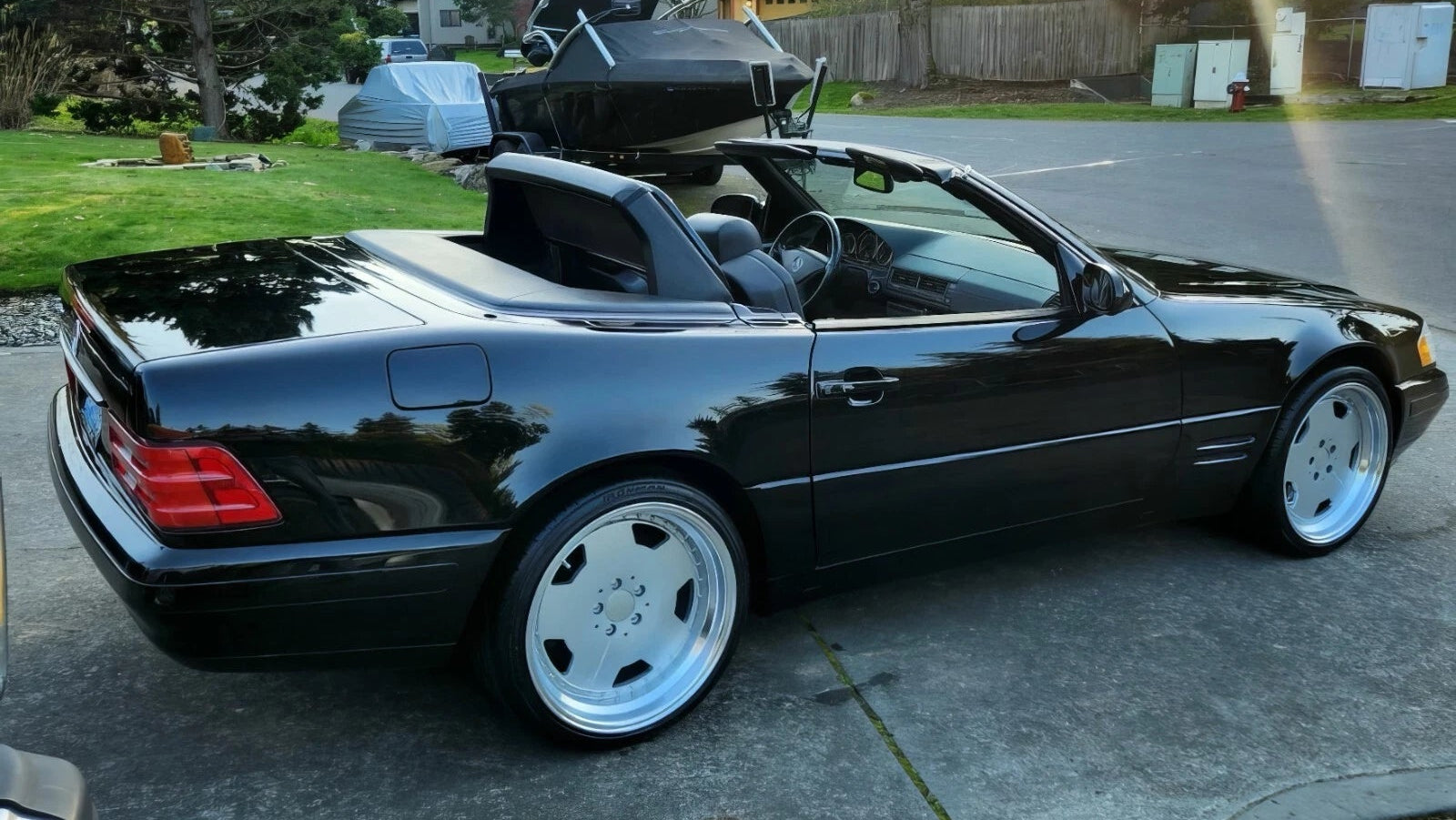 A black convertible sports car parked on a residential driveway with trees, houses, and a bright sun flare in the background.