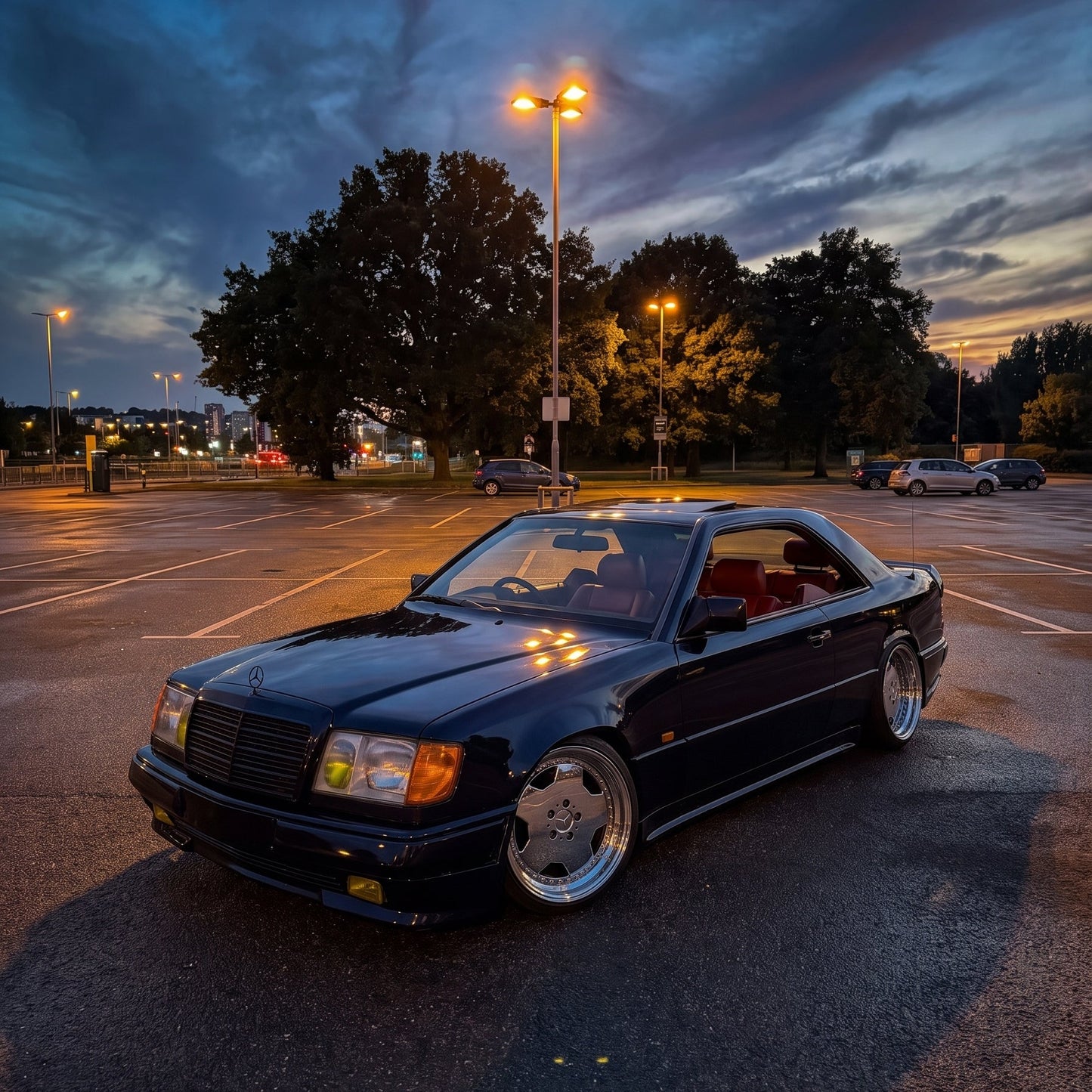 A dark blue Mercedes-Benz CLK coupe parked in an outdoor parking lot at dusk, with streetlights on and trees in the background. The car has silver multi-spoke wheels and a lowered stance.